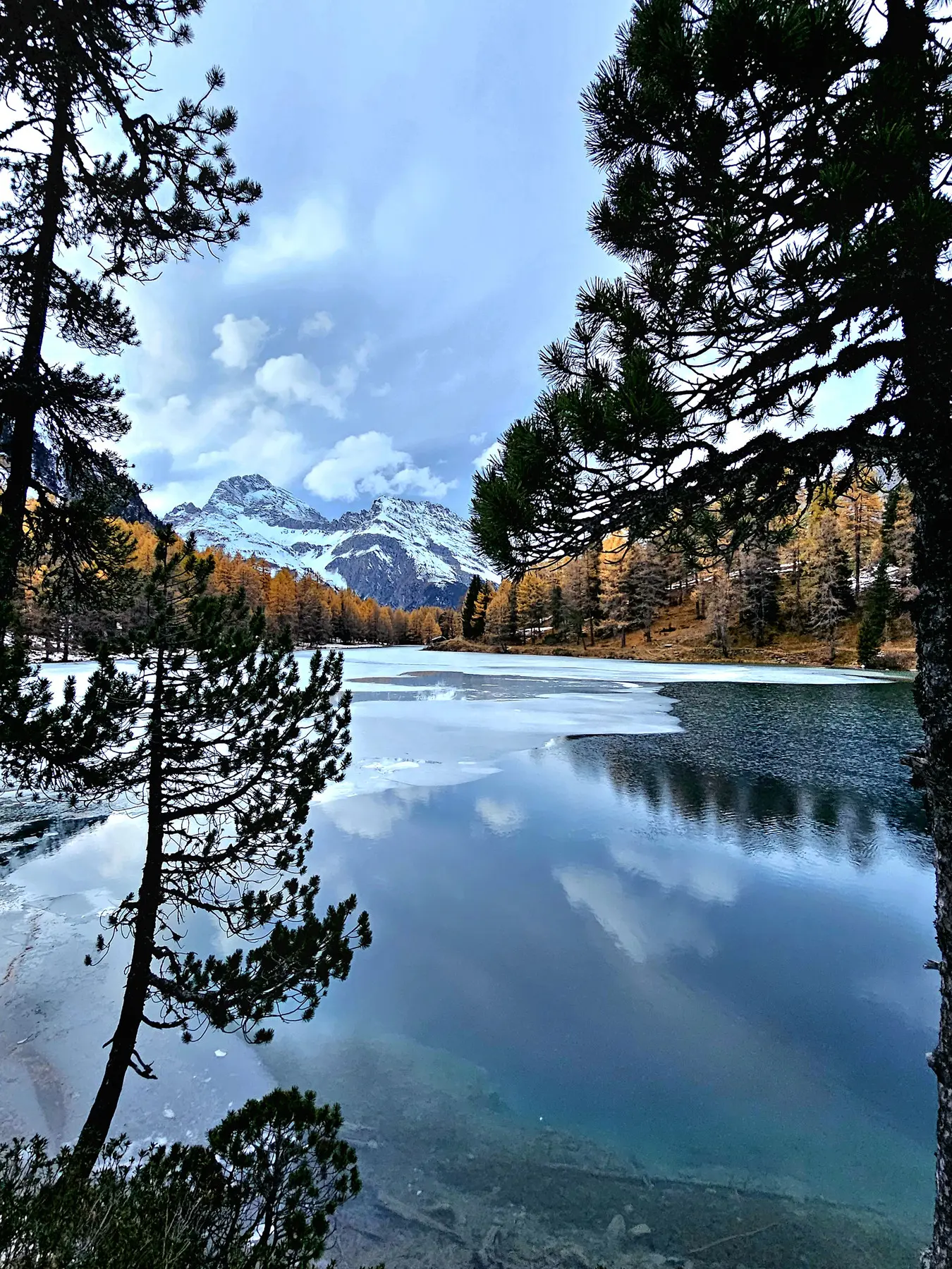 Un lago tranquillo circondato da alberi, con montagne innevate sullo sfondo e nuvole nel cielo. La superficie dell'acqua riflette il paesaggio circostante, creando un'atmosfera serena.