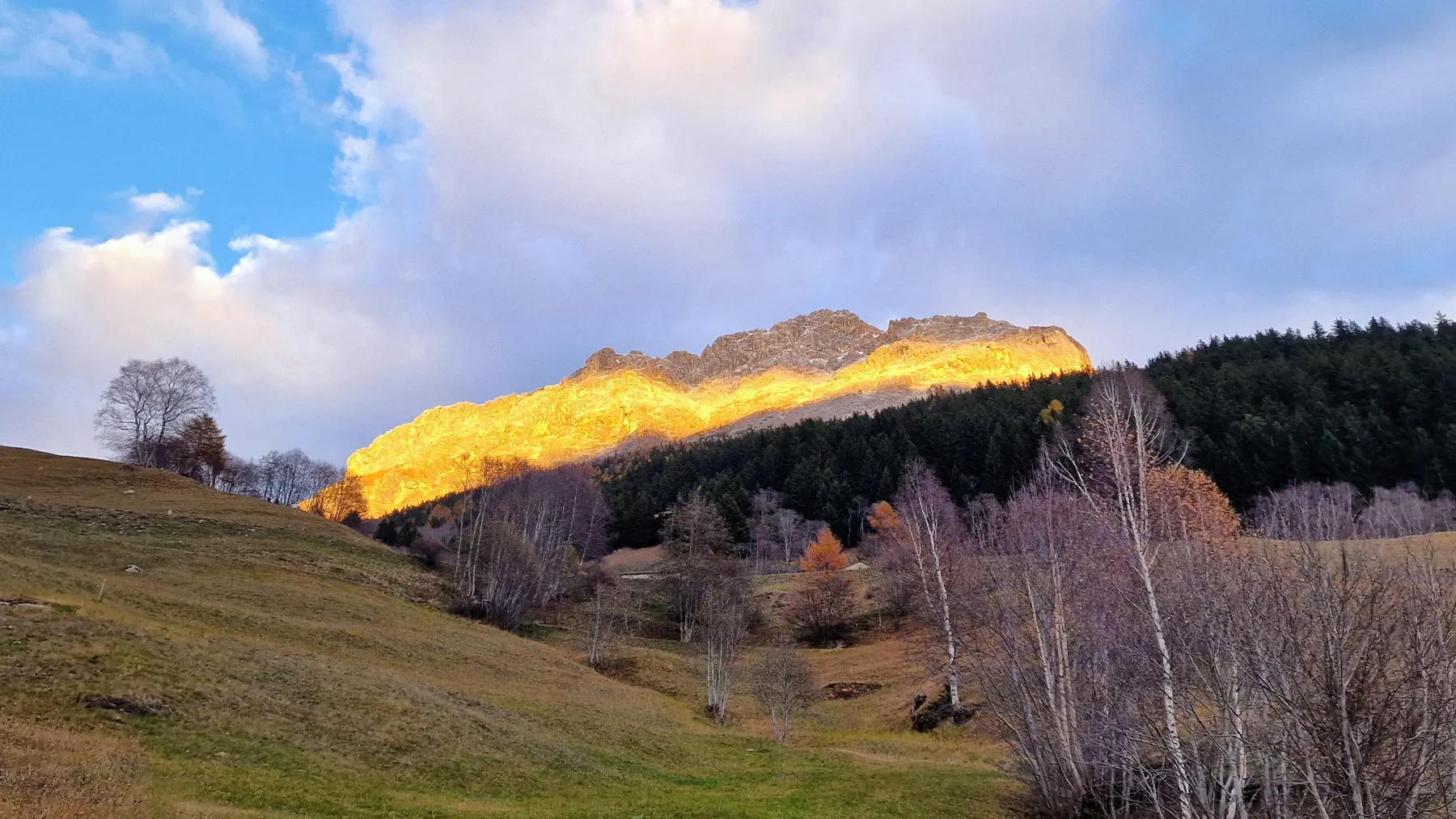 La luce del tramonto illumina la cima di una montagna, circondata da alberi e prati verdi. Il cielo è parzialmente nuvoloso, creando un contrasto suggestivo.