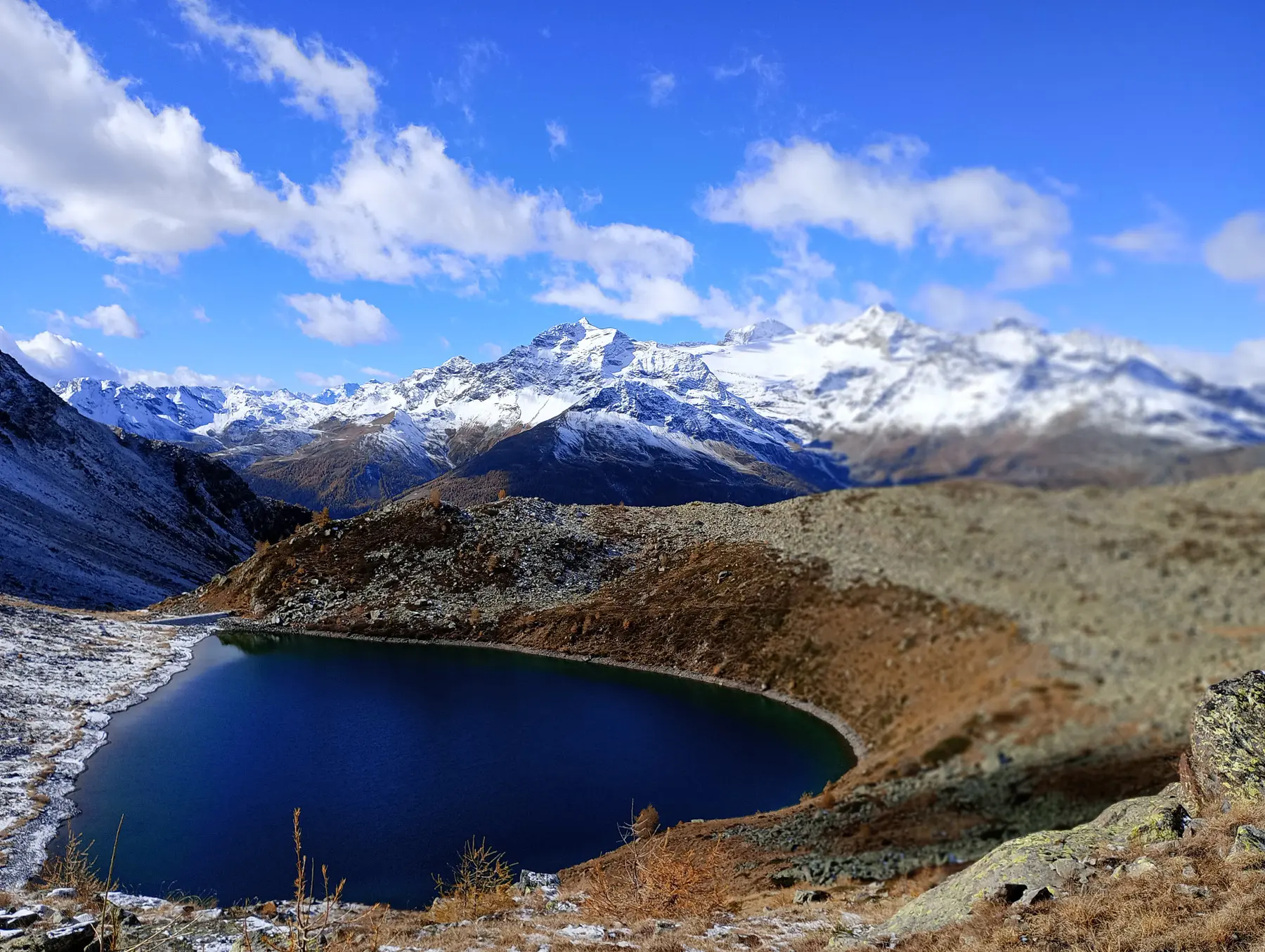 Un lago montano circondato da un paesaggio di montagne maestose, con picchi innevati e un cielo blu punteggiato di nuvole. La vegetazione secca si estende attorno al lago, creando un contrasto con l'acqua blu profonda.