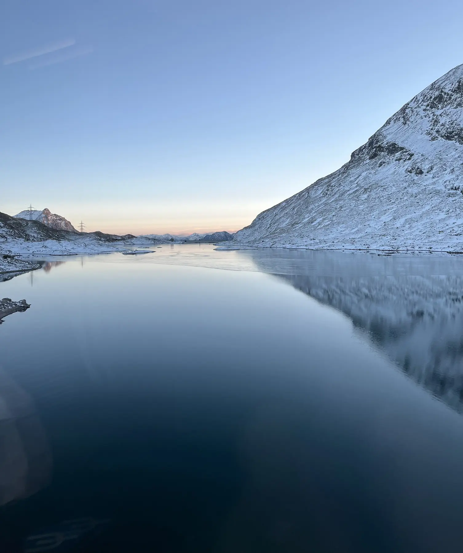 Veduta serena di un lago ghiacciato, circondato da montagne innevate al tramonto. Il cielo è di un blu tenue, riflettendo la calma dell'acqua.