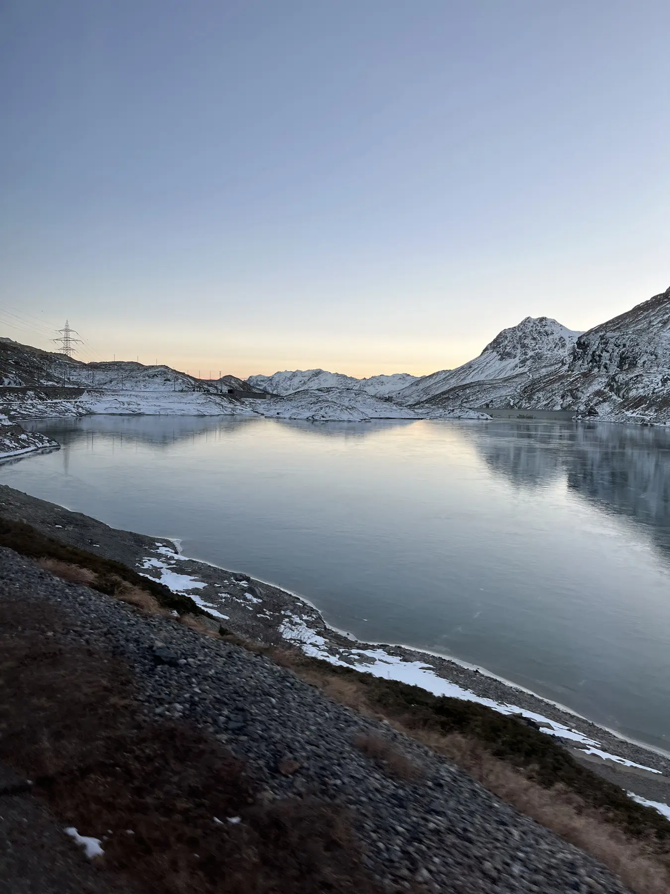 Lago montano con acqua calma e riflessi, circondato da montagne innevate sotto un cielo al tramonto.