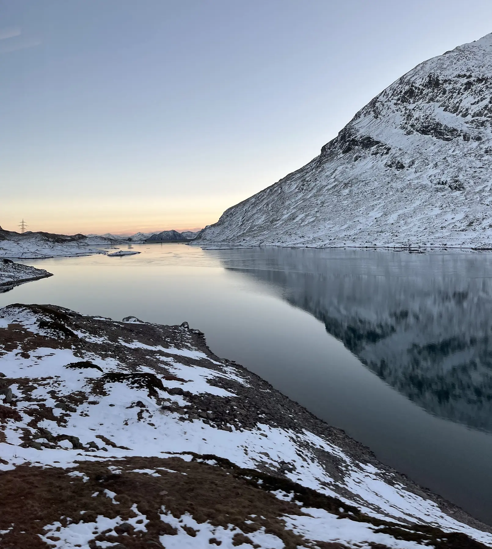 Paesaggio montano con un lago tranquillo riflettente, circondato da pendii innevati. Il cielo è chiaro con tonalità di blu e arancione al tramonto.