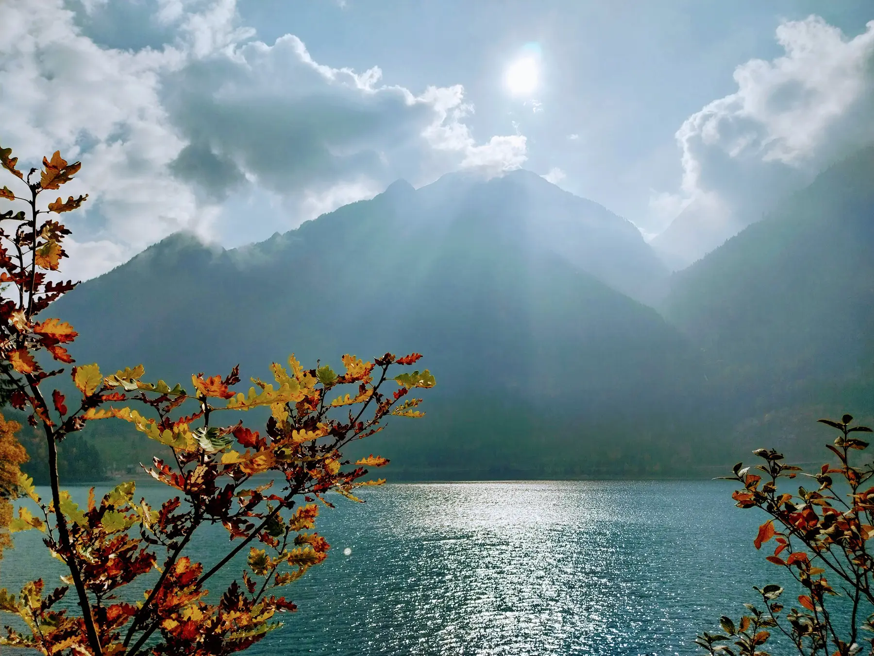 Luce solare che si riflette su un lago circondato da montagne, con foglie autunnali in primo piano.