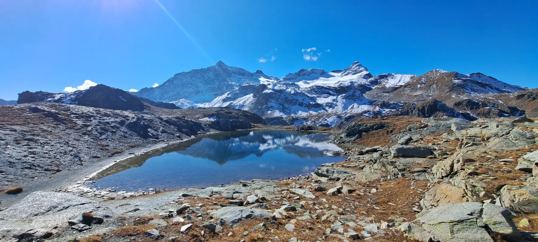 Un lago alpino segnato da rocce e prati, circondato da montagne innevate sotto un cielo blu limpido. La superficie dell'acqua riflette le cime montuose.