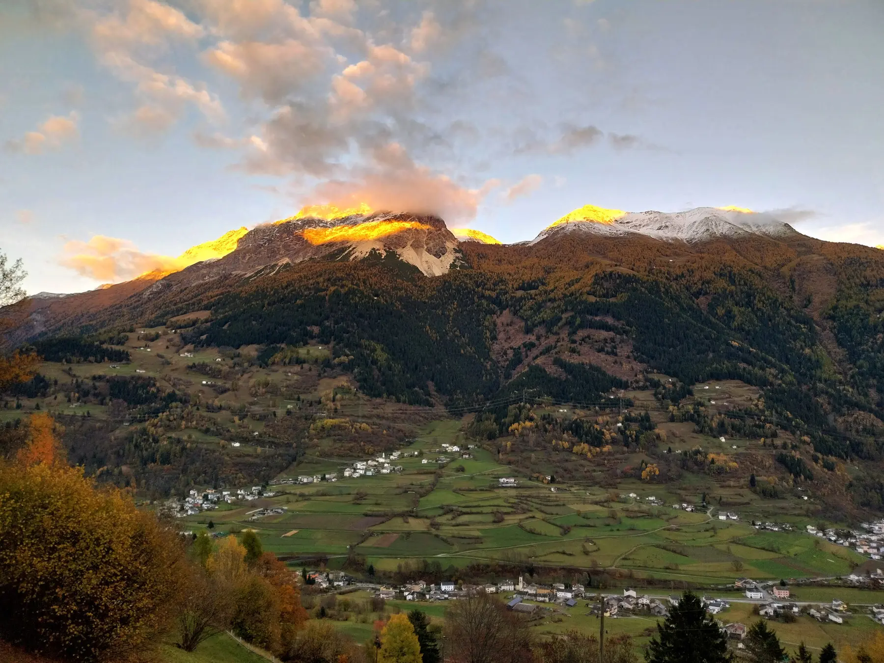 Vista panoramica di montagne al tramonto, con cime innevate illuminate dalla luce dorata. Sotto, una vallata verde con villaggi bianchi e alberi autunnali.