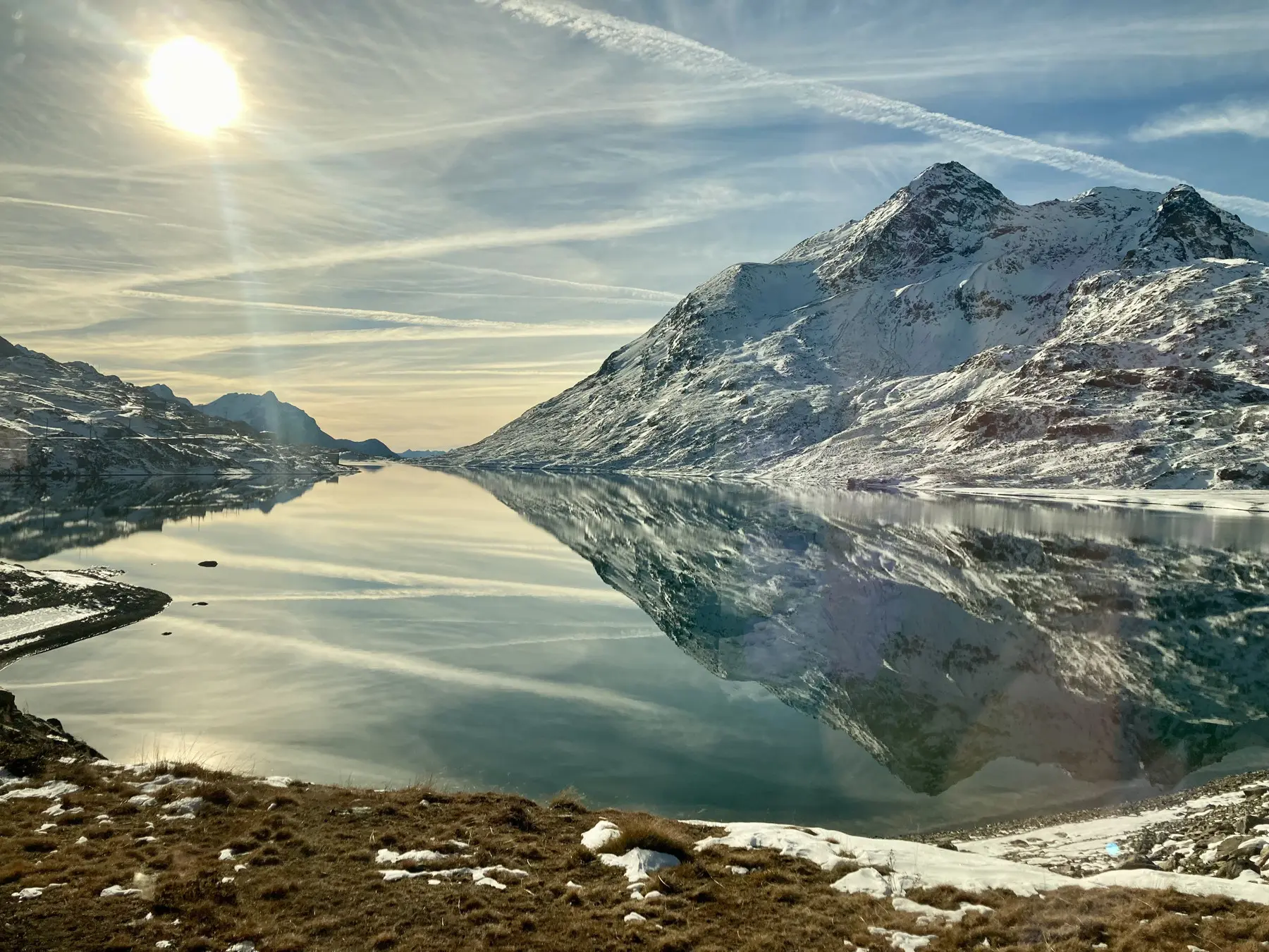 Una vista panoramica di un lago alpino tranquillo, circondato da montagne innevate. Il sole splende nel cielo blu, creando riflessi sul lago, mentre il paesaggio si riflette in maniera perfetta nell'acqua calmo.