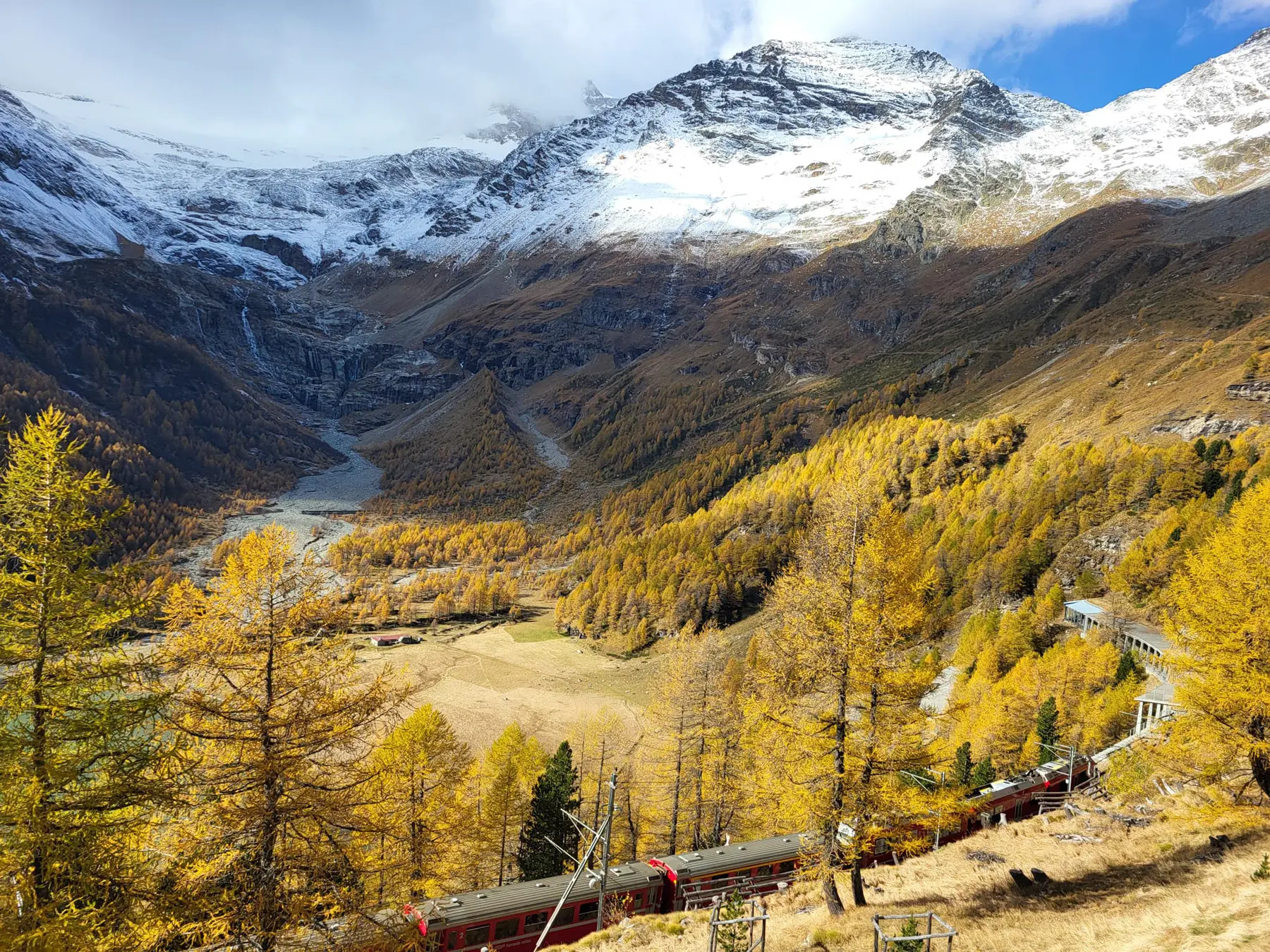 Treno rossonero si snoda in un paesaggio montano autunnale, circondato da alberi dorati e montagne innevate. Un raggio di sole illumina la valle sottostante.