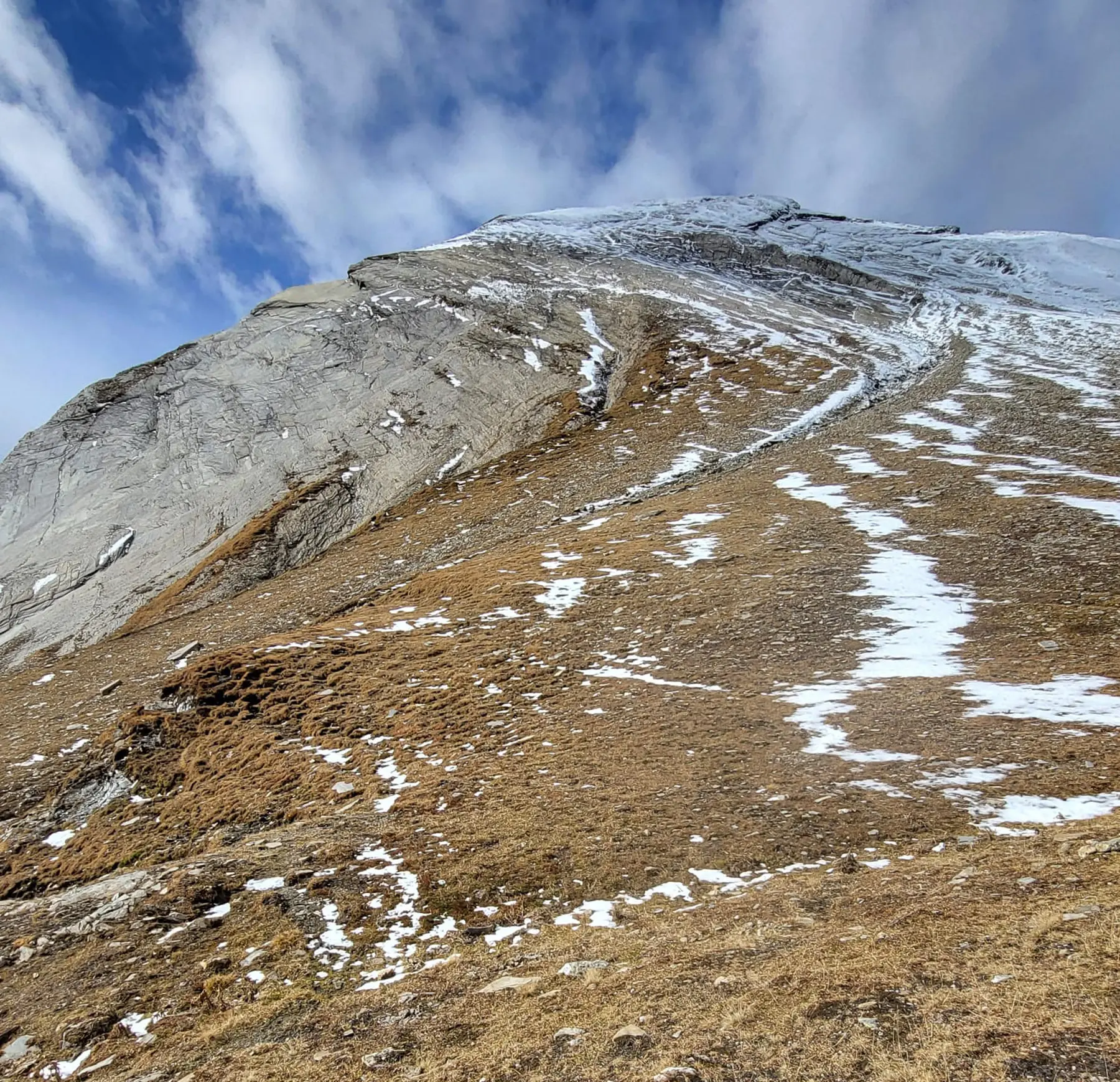 Un paesaggio montano con una grande parete rocciosa e un terreno erboso in parte coperto di neve. Il cielo è parzialmente nuvoloso, creando un contrasto tra le nuvole e la roccia.