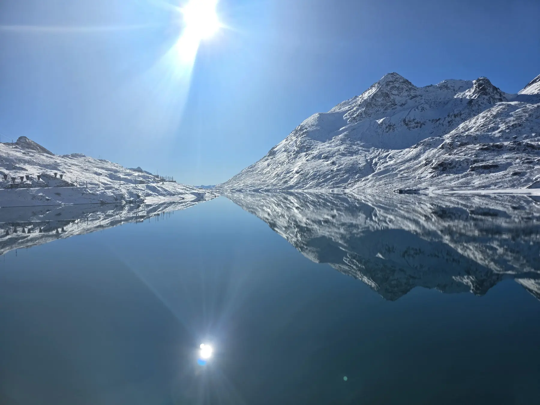 Un paesaggio alpino sereno con un lago calmo che riflette le montagne coperte di neve e il cielo blu, illuminato da un sole splendente.