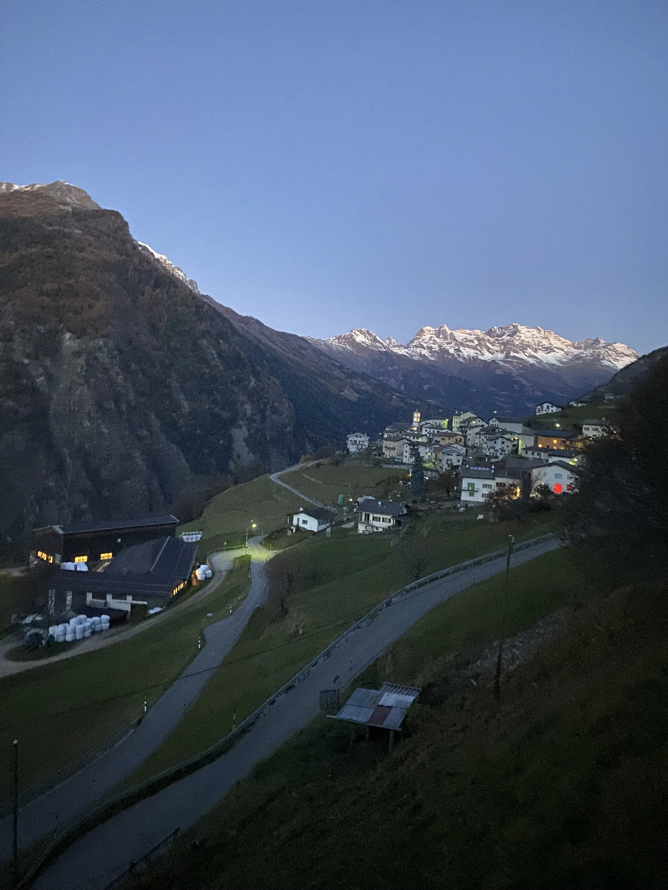 Paesaggio montano al crepuscolo, con case illuminate che si arrampicano lungo una valle. Montagne innevate sullo sfondo e una strada che si snoda tra gli edifici.
