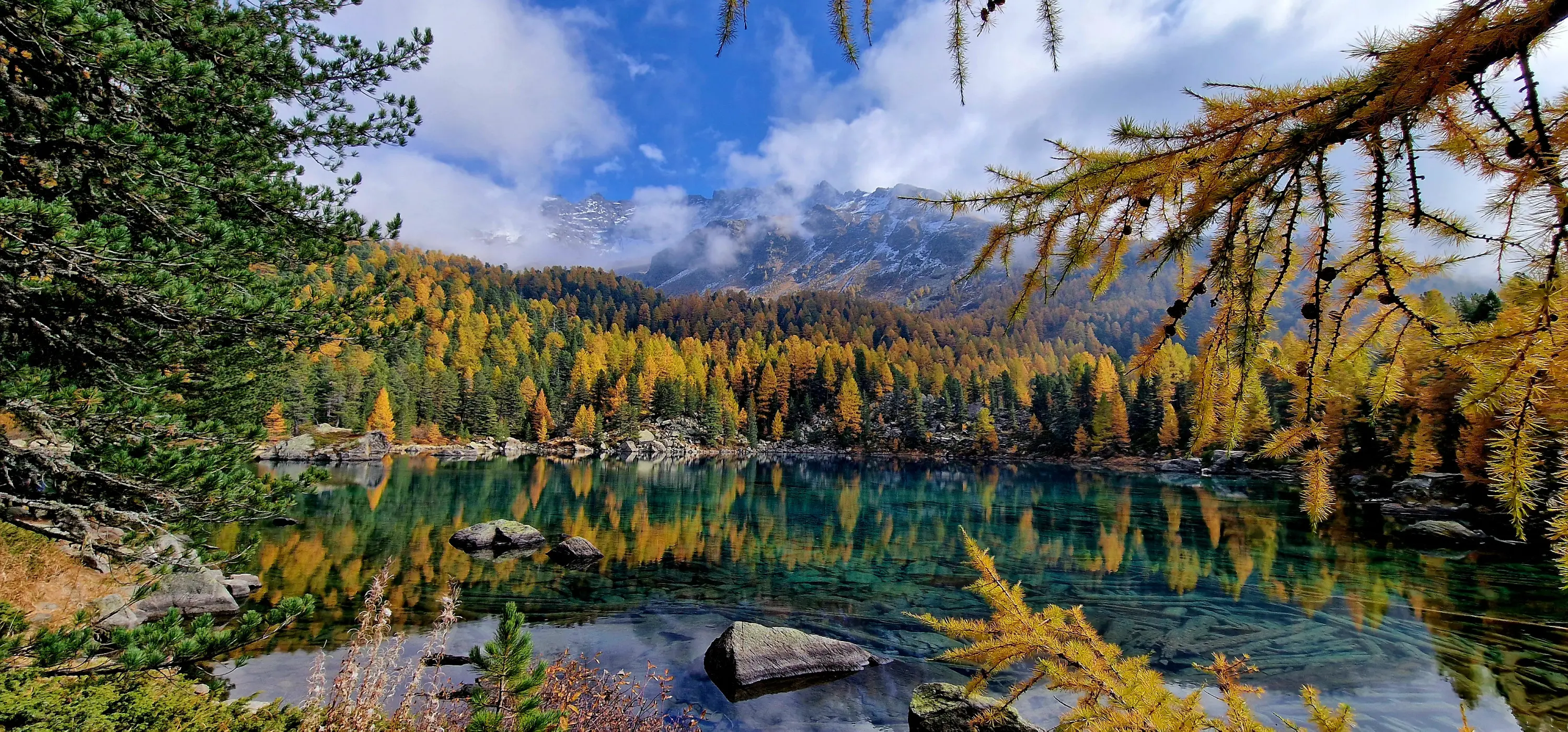 Un lago cristallino circondato da alberi autunnali dai colori vivaci, riflettendosi nell'acqua. Sullo sfondo si ergono montagne coperte di nuvole.
