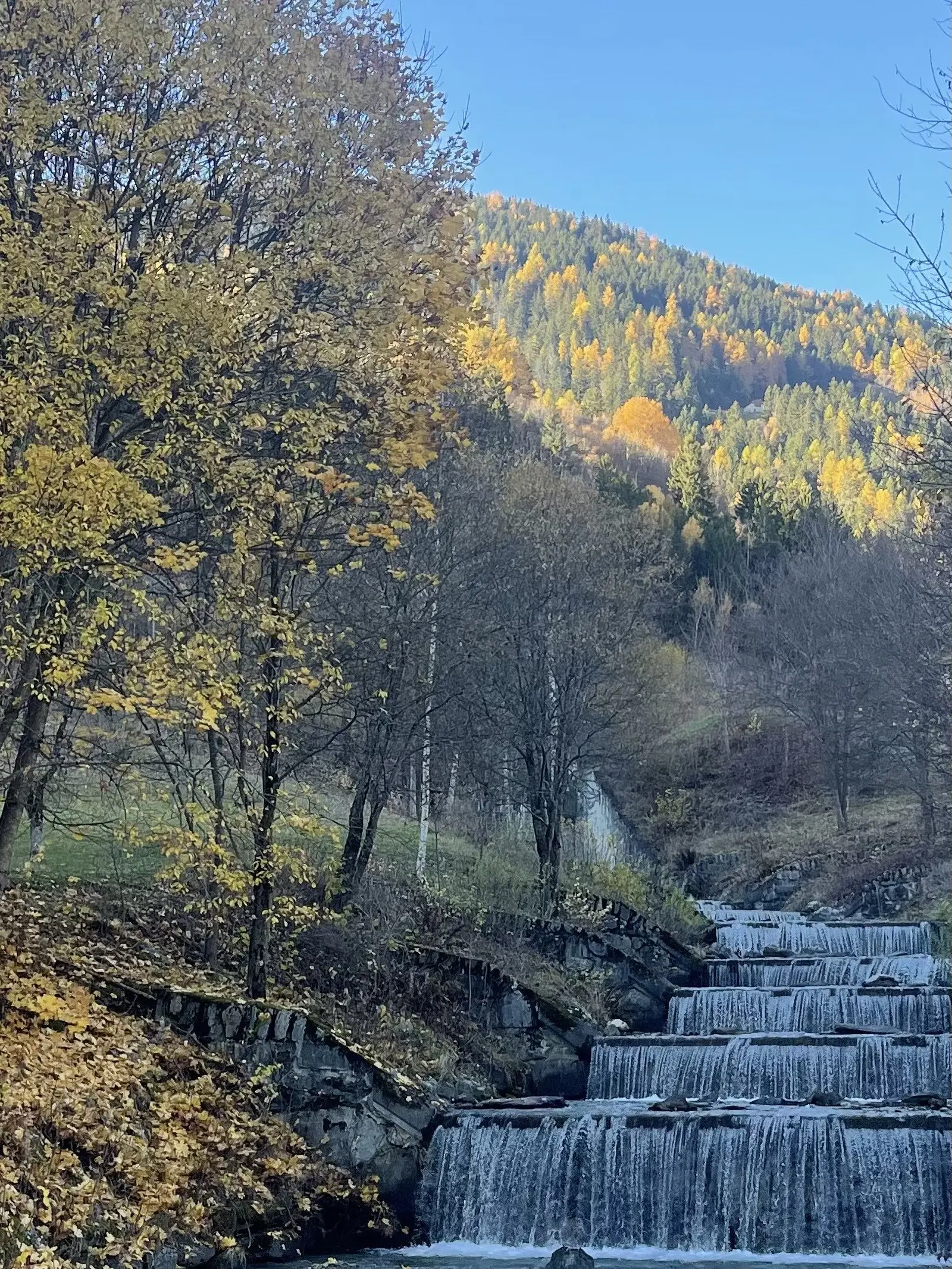Un paesaggio montano con una cascata che scorre su più livelli, circondata da alberi dai colori autunnali e un cielo blu sopra la montagna.