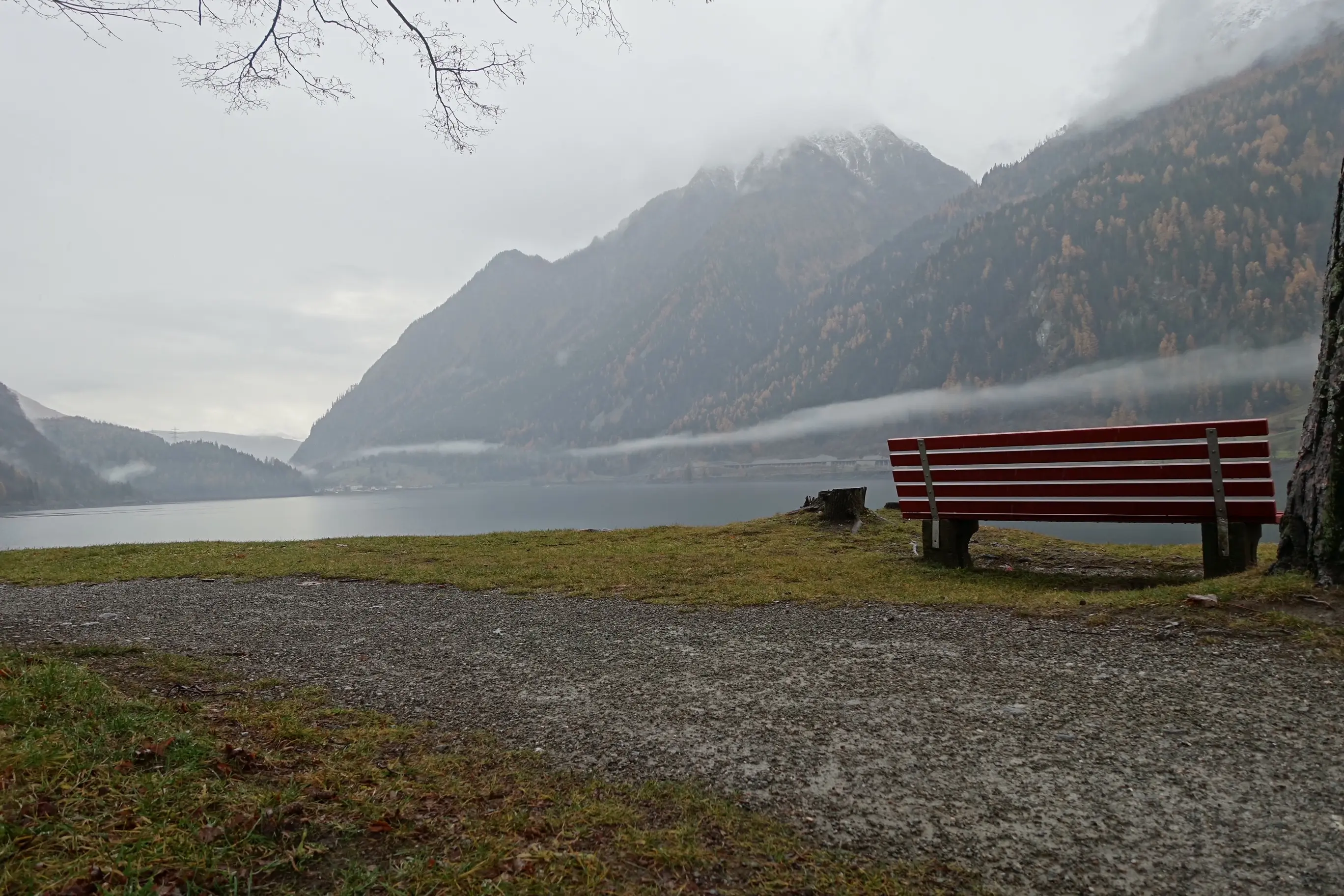 Una panchina rossa su un sentiero vicino a un lago circondato da montagne. La scena è avvolta nella nebbia con alberi autunnali dalle foglie gialle.