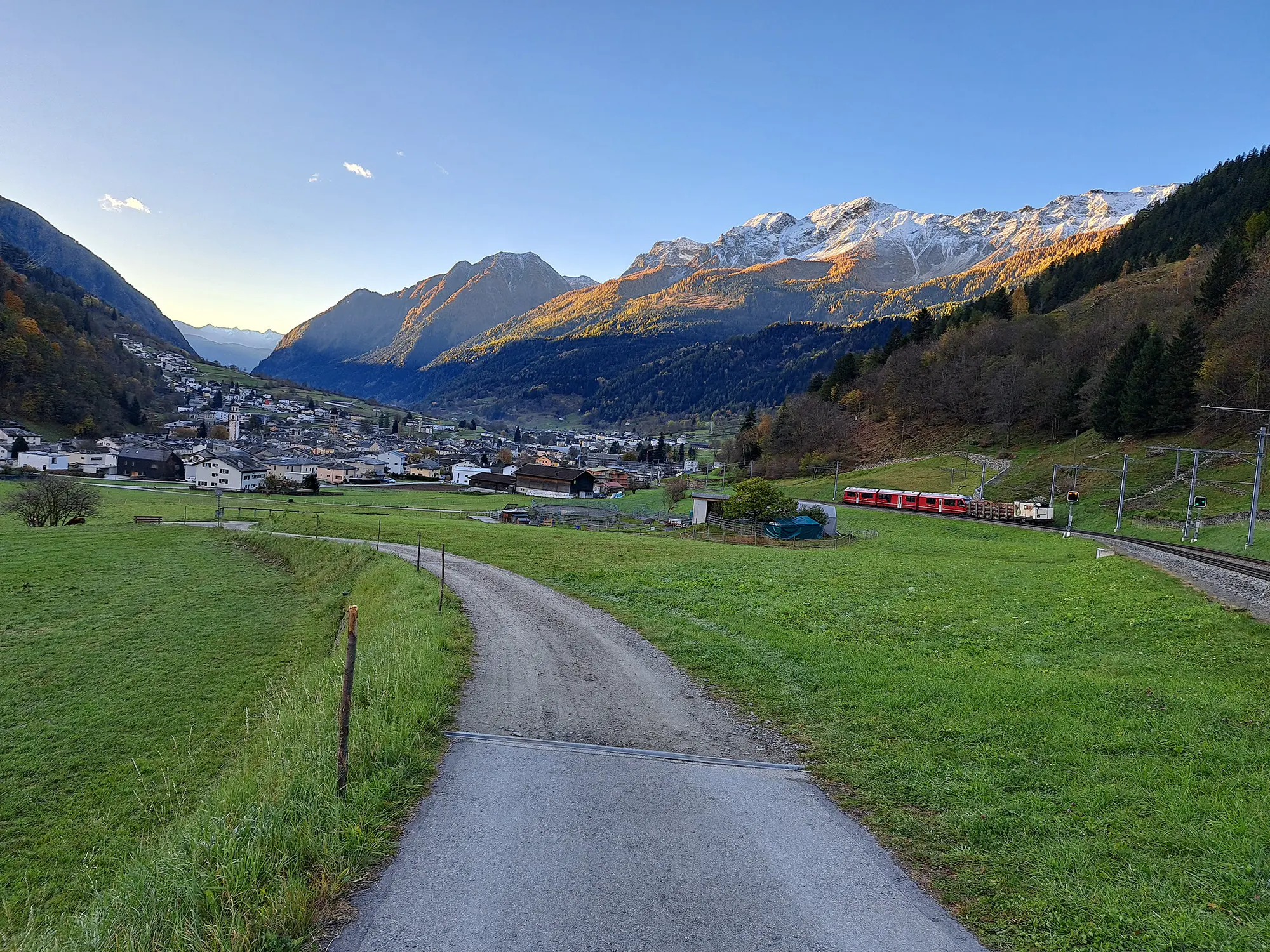Una valle verde con un sentiero che porta a un paese circondato da montagne. Le cime innevate delle montagne brillano sotto il sole. Un treno rosso attraversa la scena vicino ai binari.