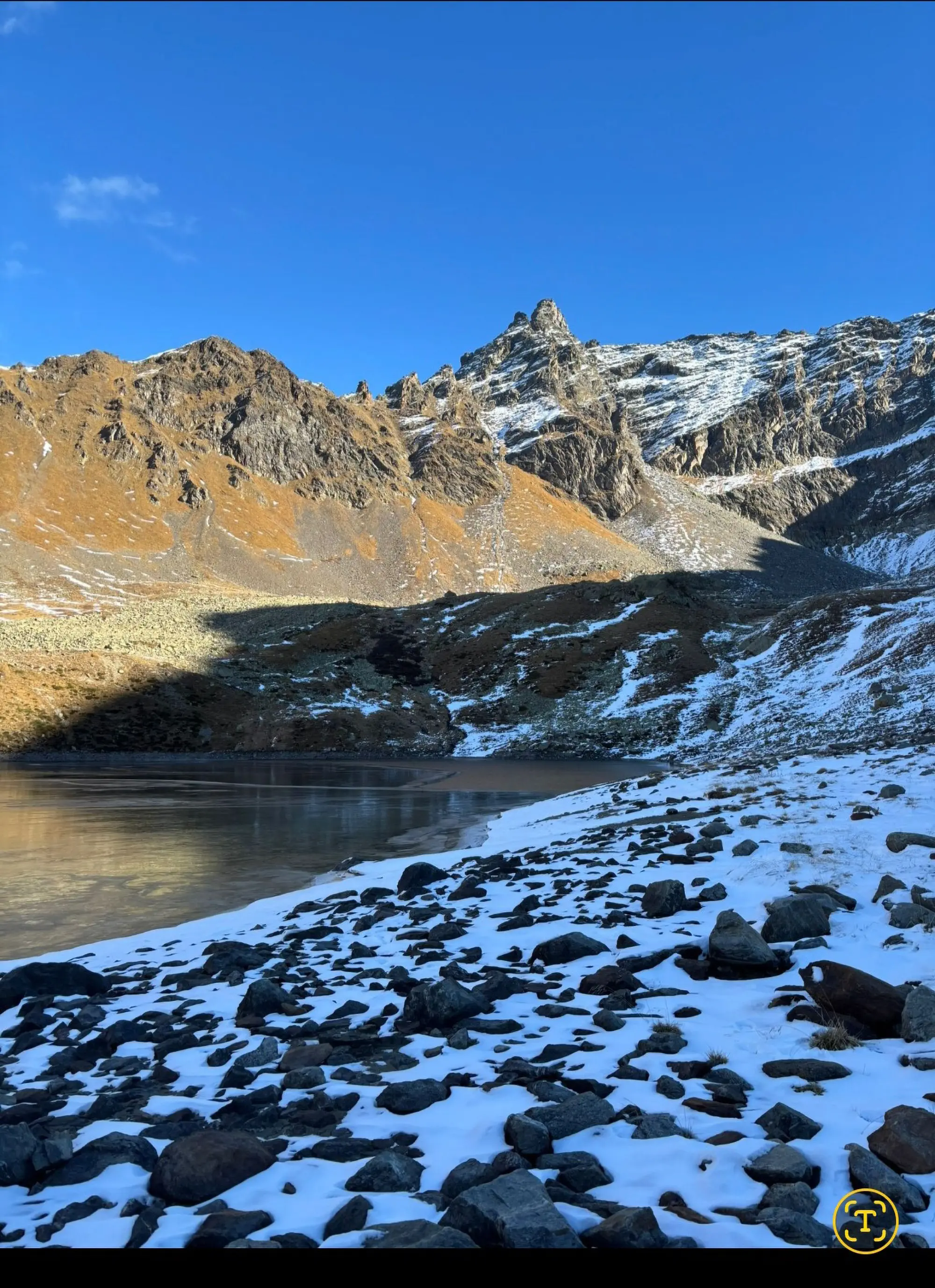 Paesaggio montano con vette rocciose, prati di erba secca e rimanenze di neve. Un lago ghiacciato si trova in primo piano, con un cielo azzurro e chiaro sullo sfondo.