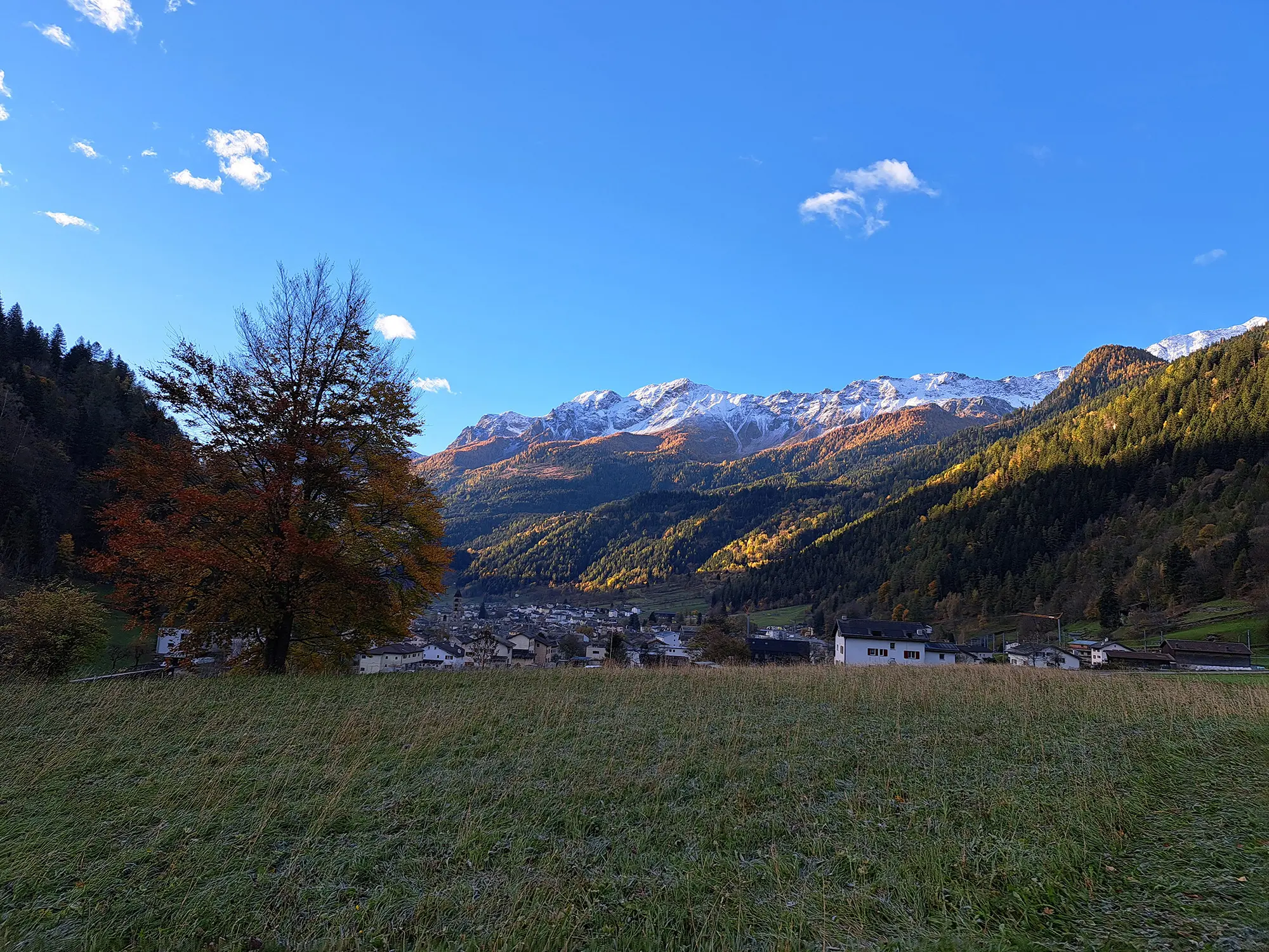 Panorama di un villaggio montano circondato da alberi autunnali e cime innevate. Il cielo è blu con alcune nuvole bianche.