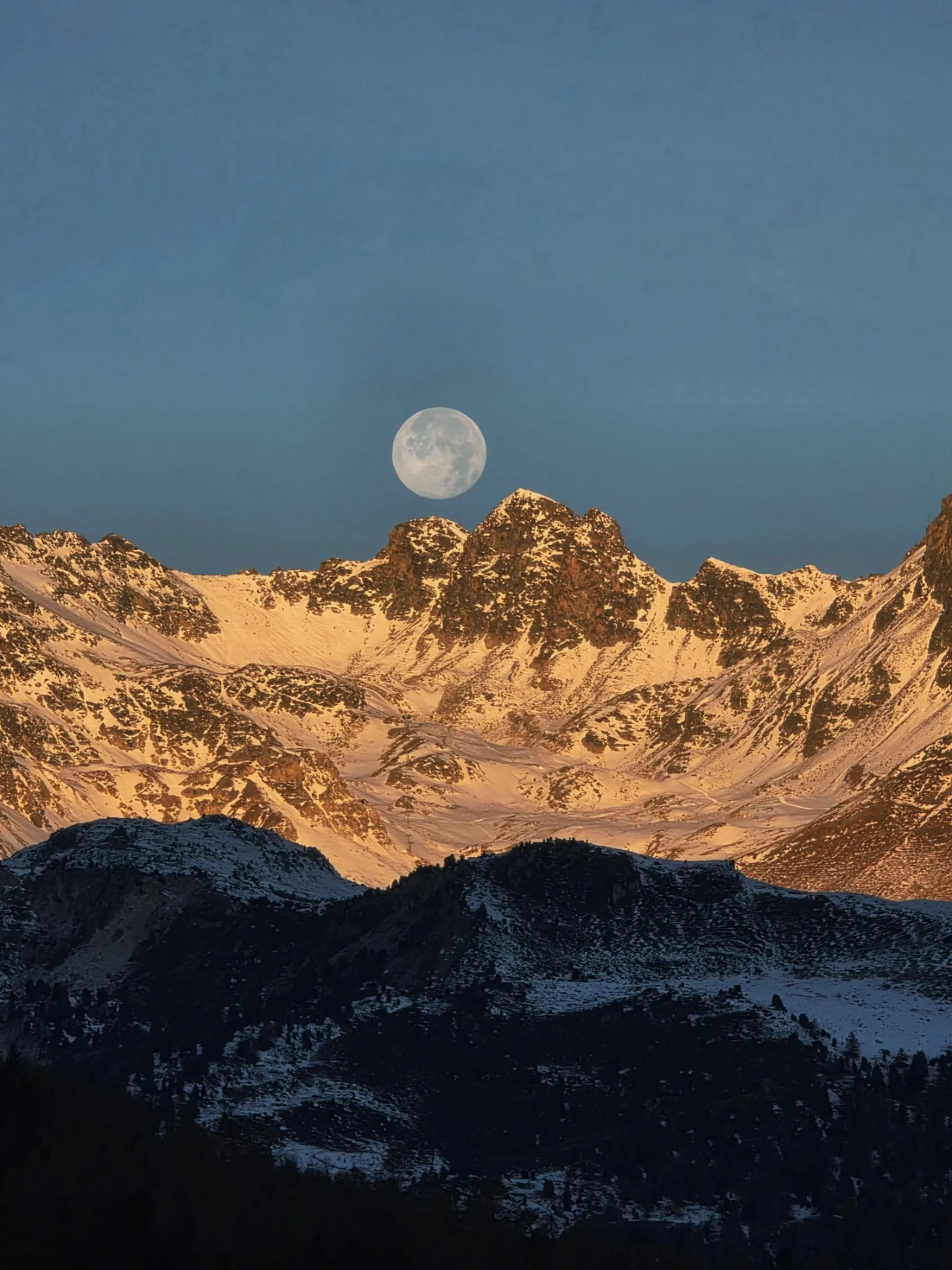 Una grande luna piena si erge sopra le montagne innevate, illuminate dalla luce dorata del tramonto. Le cime rocciose si stagliano contro un cielo blu, creando un'atmosfera serena e suggestiva.